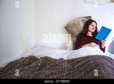 une jeune femme lit allongé dans un lit avec un livre bleu Banque D'Images
