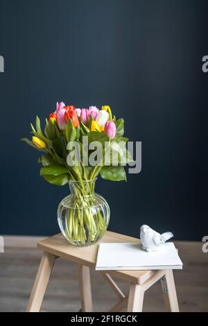 Un bouquet de tulipes multicolores dans un vase transparent sur la chaise haute avec marche sur fond bleu foncé. Ordinateur portable blanc. Banque D'Images