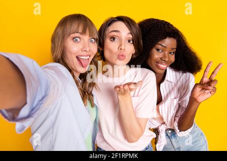 Photo de Nice optimiste trois dames ne selfie coup baiser montrez le v-sign tongue out portez une chemise blanche isolée sur brillant arrière-plan de couleur jaune Banque D'Images