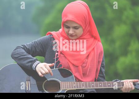 Portreit de la jeune fille musulmane préateuse portant le hijab essayant de jouer guitare extérieur Banque D'Images