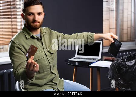 Nouvelles technologies. Un jeune homme jette un portefeuille avec de l'argent dans un sac à ordures et tient une carte bancaire à l'appareil photo. Paiement en ligne. Paiement sans espèces. Banque D'Images