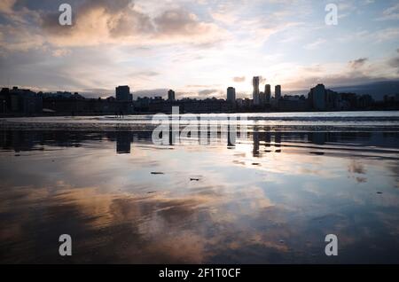 Les nuages roses réfléchissent sur le sable humide sur une plage appelée Playa Bristol, Mar del Plata, Argentine. Silhouette de plusieurs surfeurs et du centre-ville Banque D'Images