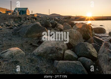 Heure d'or dans la communauté inuite de Qikiqtarjuaq, île Broughton, Nunavut, Canada. Grands rochers avec maisons en arrière-plan et soleil à l'horizon. Banque D'Images