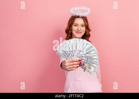 Portrait d'une adolescente heureuse à cheveux bouclés à capuche avec halo sur la tête montrant beaucoup de billets de banque en dollars et souriant, charité. Prise de vue en studio en intérieur isola Banque D'Images