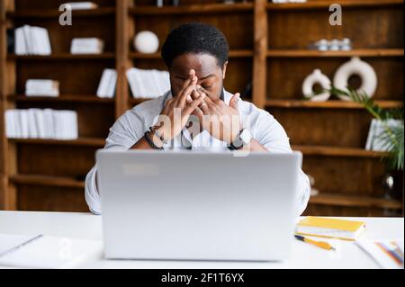 Surpassé le travail en ligne sur un ordinateur portable afro-américain homme sentant la tension des yeux, burnout, biracial mâle indépendant massage pont du nez assis sur le lieu de travail dans un espace de bureau moderne Banque D'Images