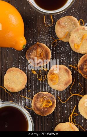 Plats de biscuits faits maison, thé noir, citron, zeste de citron et glaçage au sucre sur une planche en bois sombre nervurée vue au plafond Banque D'Images