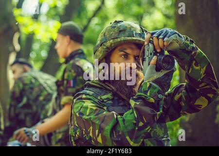 Journaliste soldat avec un visage sérieux dans des munitions et un casque tenant la caméra dans ses mains sur le fond des soldats dedans forêt Banque D'Images