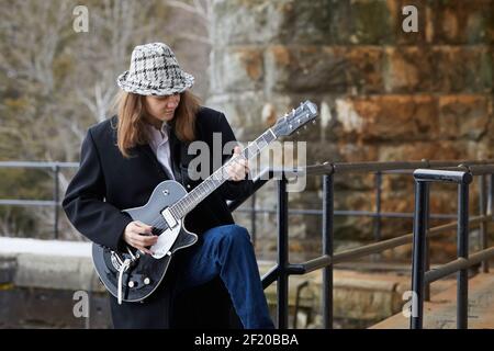 Jeune musicien masculin en manteau et chapeau debout sur les escaliers et jouer de la guitare électrique à l'extérieur du vieux bâtiment Banque D'Images