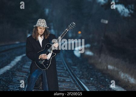 Personne portant un long manteau et chapeau à motifs joue de la guitare électrique tout en se tenant debout sur les voies ferrées à l'extérieur. Banque D'Images