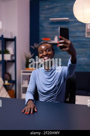 Femme à la peau sombre et gaie qui prend un autoportrait en utilisant un smartphone tard dans la nuit de la maison. Travailleur indépendant utilisant un réseau de technologie moderne sans fil faisant des heures supplémentaires. Banque D'Images