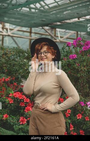 Une belle fille de taille plus en lunettes et chapeau souriant parmi les plantes vertes et les fleurs. Banque D'Images