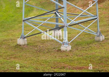 Partie inférieure d'un grand support électrique en acier sur fond d'herbe verte, structure métallique attachée à une base en béton dans le sol, haute tension a Banque D'Images