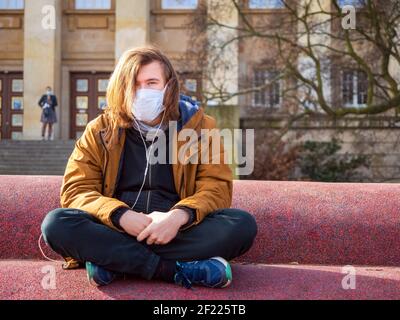 Le jeune homme triste porte un masque médical de protection pour se protéger du coronavirus pandémique en plein air et assis seul dans une ville et écoutez de la musique Banque D'Images