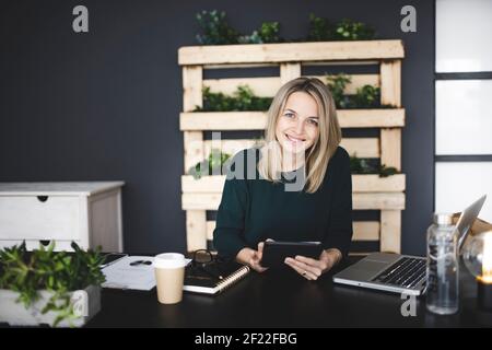 jolie jeune femme blonde est assise dans un bureau moderne et durable avec beaucoup de plantes écologiques vertes et travaille sur sa tablette