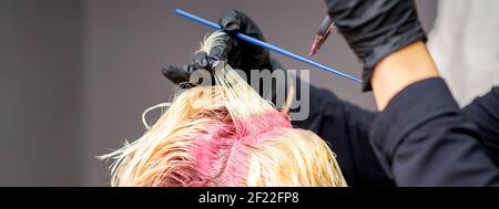 Gros plan sur les mains du coiffeur en appliquant du colorant rose sur celles de la femme cheveux blonds dans un salon de coiffure Banque D'Images