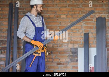 Homme de construction confiant portant une combinaison et un casque dur regardant concentré tout en tenant un goujon en métal pour cloison sèche, travaillant sur la construction de maison. Bâtiment, profession, concept de sécurité Banque D'Images