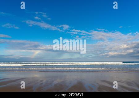 Une plage de sable avec de grandes vagues et un petit arc-en-ciel dans un ciel expressif Banque D'Images