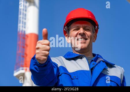 Un travailleur de la centrale électrique qui se donne contre les cheminées industrielles et le fond Blue Sky. Travailleur dans un équipement de protection individuelle regardant la caméra. Banque D'Images