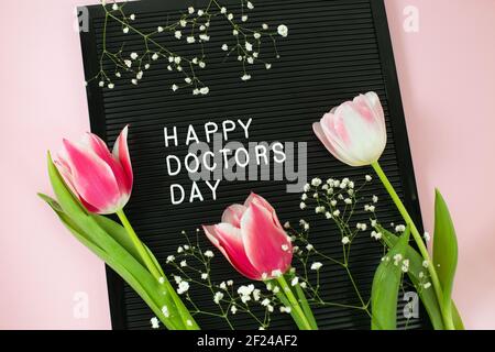 Carton noir avec lettres en plastique blanc avec citation Happy Doctor's Day et bouquet de tulipes roses sur fond rose. Journée nationale des médecins. Bonne journée infirmière. plat Banque D'Images