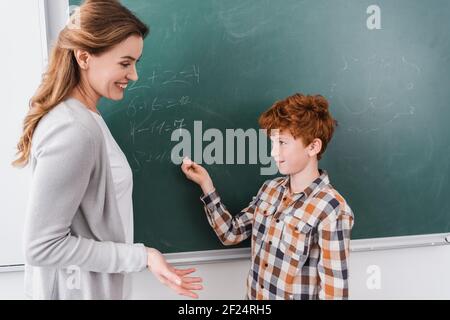 professeur souriant pointant avec la main près de la tête rouge élève écrivant dessus tableau noir Banque D'Images