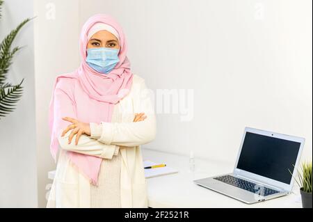 Jeune femme d'affaires arabe musulmane confiante dans le hijab et masque de protection debout avec les bras repliés sur le lieu de travail avec un bureau et un ordinateur portable, regardant la caméra, la sécurité au travail concept Banque D'Images