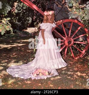 1980s vue d'archive historique de 1989 jeune modèle libéré mariée posant pour la photographie à l'extérieur à côté de roue de cartwheel prop portant toute la longueur hors de l'épaule 80s robe de mariée et train en couleur photo avec bouquet et coiffe assortie image de mode d'archive de la façon dont nous étions dans les années 1900 Essex Angleterre Royaume-Uni Banque D'Images