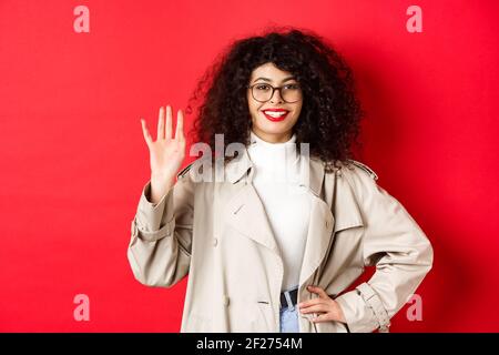 Femme européenne élégante en lunettes et en trench-coat, se lever la main et sourire, en disant bonjour, saluant quelqu'un, debout sur le bac rouge Banque D'Images