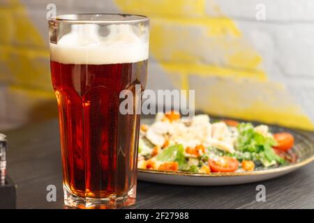 Verre de bière foncée avec mousse et salade César en plaque d'argile, sur table en bois noir Banque D'Images