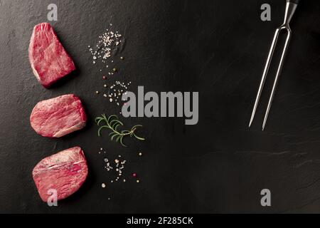 Filets de bœuf crus avec sel, poivre, romarin et une fourchette à découper sur fond noir Banque D'Images