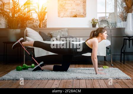 Une jeune fille de forme physique sportive forte et belle se produit des coups de pied couchés avec des bracelets de sport dans son lumineux et chambre spacieuse avec un mini Banque D'Images