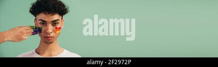 Portrait d'un jeune homme au visage peint avec un drapeau arc-en-ciel de fierté gay. Peinture à la main de couleur arc-en-ciel lgbt sur le visage d'un homme sur fond vert avec Banque D'Images