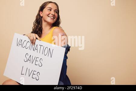 Une femme joyeuse tenant un panneau indiquant « la vaccination sauve des vies » écrit dessus. Femme avec bandage sur son bras après avoir reçu la vaccination tenant un ba Banque D'Images