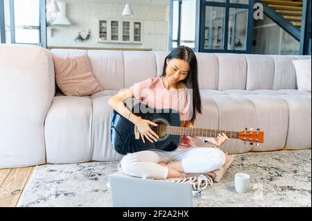 Jolie femme asiatique aux longs cheveux noirs jouant joyeusement la guitare, assise sur le sol dans le salon, donnant des cours de musique en ligne, l'enseignement, la performance en streaming dans les médias sociaux sur l'ordinateur portable Banque D'Images