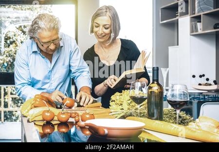 Couple senior cuisant des aliments sains et buvant du vin rouge à Cuisine maison - les gens retraités à la maison préparant le déjeuner avec livre de recettes Banque D'Images