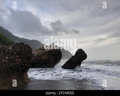 Un pêcheur assis sur une formation rocheuse tandis que de grandes vagues coulent et se déversent rapidement contre la falaise et la large plage de sable à Parangtritis, Java. Banque D'Images