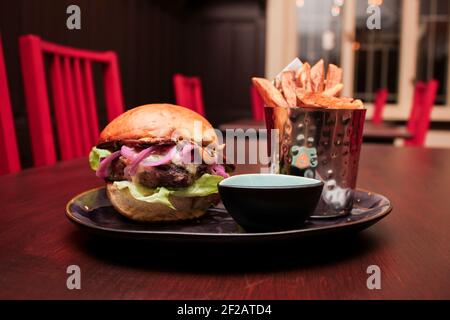 Hamburger de bœuf juteux à l'oignon rouge, frites dans un seau en métal brillant et sauce sur une assiette faite à la main à l'intérieur sombre du restaurant. Banque D'Images