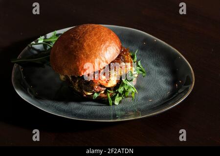 Hamburger de bœuf juteux avec oignon caramélisé, fromage et rocola sur un petit pain maison. Hamburger servi sur une assiette grise artisanale vintage sur la table marron foncé. Banque D'Images