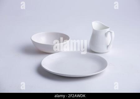 Articles de table blancs, différentes formes d'une plaque en céramique blanche vide, bol et théière disposés sur un fond blanc texturé. Banque D'Images