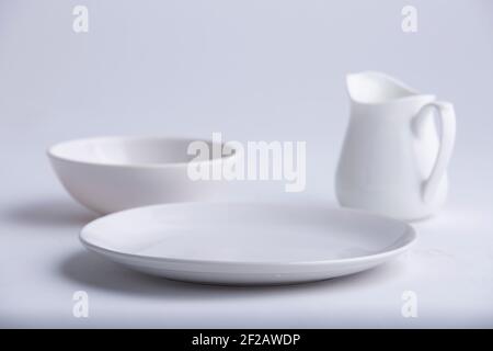 Articles de table blancs, différentes formes d'une plaque en céramique blanche vide, bol et théière disposés sur un fond blanc texturé. Banque D'Images