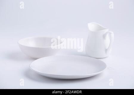 Articles de table blancs, différentes formes d'une plaque en céramique blanche vide, bol et théière disposés sur un fond blanc texturé. Banque D'Images