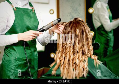 Vue arrière des mains du coiffeur féminin qui froulent les cheveux des femmes avec fer à friser dans un salon de coiffure Banque D'Images