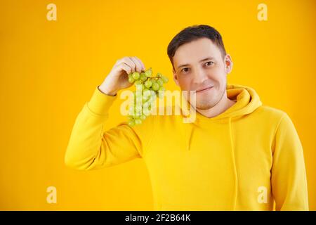 Portrait d'un jeune homme avec des raisins verts sur fond jaune. Gros plan d'un beau gars à capuchon jaune tient une bande de raisin Banque D'Images