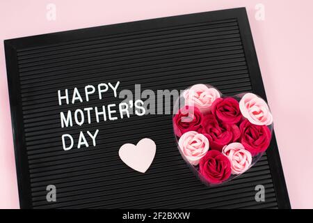 Lettrage noir avec lettres en plastique blanc avec citation Happy Mothers day et savon de fleur, coeur sur fond rose. Banque D'Images