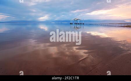 Calme mer morte surface dans le matin, ciel rose et bleu au-dessus, photo grand angle, détail de mise au point sur sable premier plan, arrière-plan d'abri de soleil flou, typi Banque D'Images