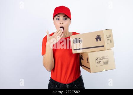 Jeune belle femme portant l'uniforme de messagerie tenant des colis de livraison couvrant la bouche avec la main, choqué et peur pour l'erreur. Expression surprise Banque D'Images