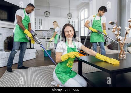 Jolie femme caucasienne souriante dans un tablier vert et des gants jaunes, essuyant la table avec un chiffon en microfibre et des détergents et souriant à l'appareil photo. Nettoyage Banque D'Images