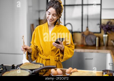 Une femme heureuse vêtue de peignoir prépare des crêpes pour le petit déjeuner, regarde au téléphone tout en se tenant près de la plaque de cuisson dans la cuisine moderne à la maison Banque D'Images