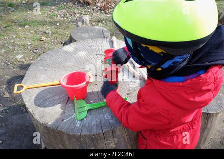 Petit garçon en manteau rouge et casque de vélo jaune jouant avec des conteneurs de jouets en sable et en plastique sur une bûche en bois en plein air dans la banlieue de Budapest, Hongrie Banque D'Images