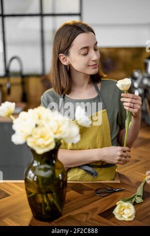 La jeune femme au foyer en tablier vert décorera l'intérieur de la maison avec un bouquet de tulipes fraîches. Fleuriste avec fleurs sur la cuisine à la maison. Tenir la fleur à la main près du vase. Banque D'Images