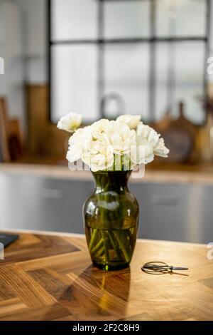 Vase avec fleurs blanches sur fond intérieur élégant de cuisine avec table en bois. Concept de confort à la maison. Bouquet de tulipes dans vase en verre vert. Banque D'Images